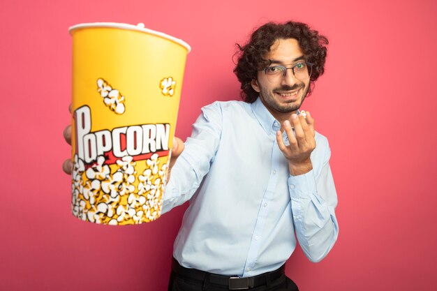 Smiling young handsome caucasian man wearing glasses looking at camera stretching out bucket of popcorn towards camera with popcorn piece in another hand isolated on crimson background with copy space