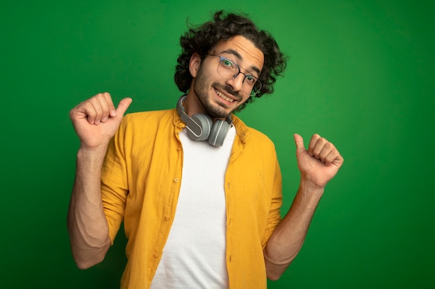 Free Photo smiling young handsome caucasian man wearing glasses with headphones around neck looking at camera pointing at himself isolated on green background