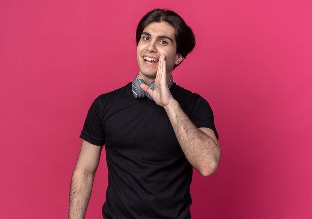 Smiling young handsome guy wearing black t-shirt and headphones around neck calling someone isolated on pink wall