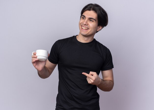 Smiling young handsome guy wearing black t-shirt holding cup of coffee points at side isolated on white wall with copy space