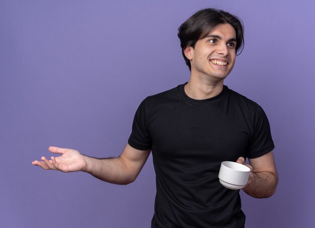 Smiling young handsome guy wearing black t-shirt holding cup of coffee spreading hand isolated on purple wall