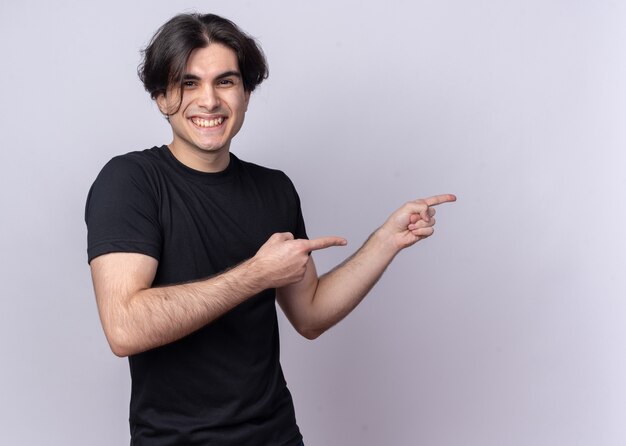 Smiling young handsome guy wearing black t-shirt points at side isolated on white wall with copy space