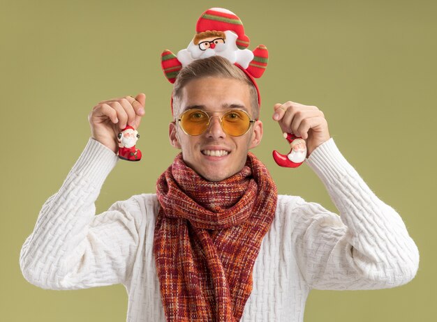 smiling young handsome guy wearing santa claus headband and scarf looking  holding santa claus christmas ornaments isolated on olive green wall