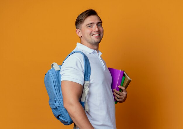 Smiling young handsome male student wearing back bag holding books isolated on orange