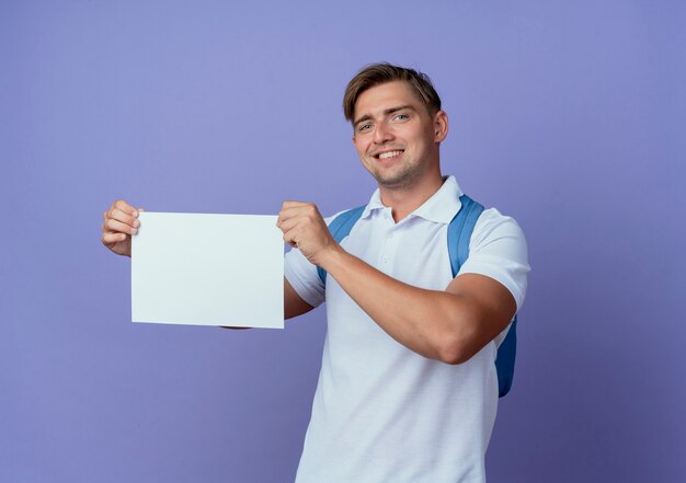 Smiling young handsome male student wearing back bag holding paper isolated on blue