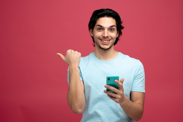 smiling young handsome man holding mobile phone looking at camera pointing to side isolated on red background with copy space