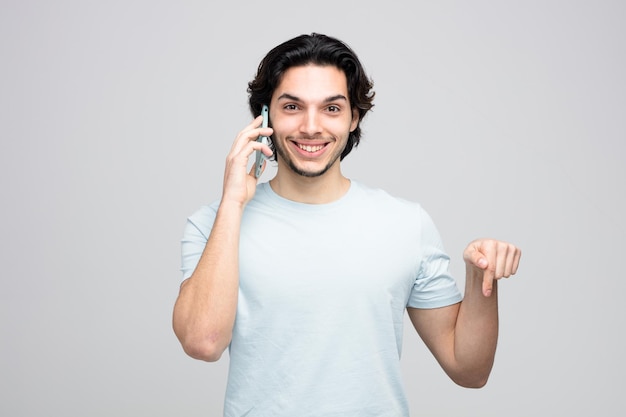 smiling young handsome man looking at camera pointing down while talking on phone isolated on white background