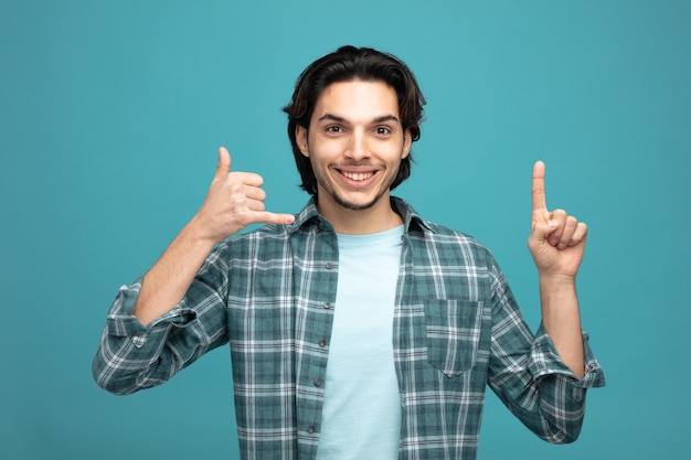 smiling young handsome man looking at camera pointing up showing cal gesture isolated on blue background