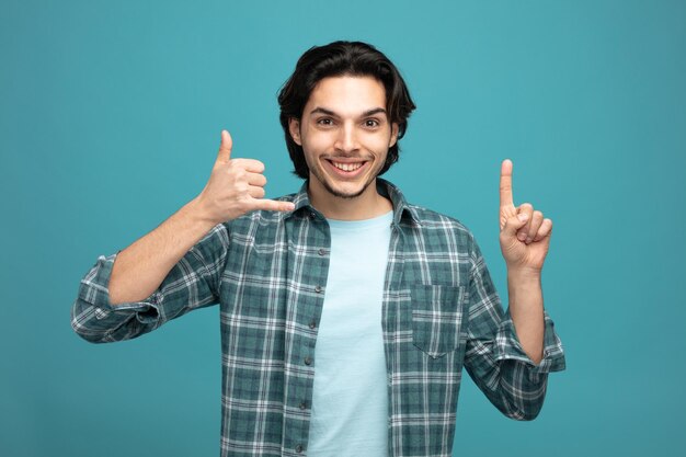 smiling young handsome man looking at camera showing call me gesture pointing up isolated on blue background