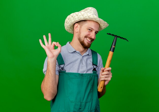 Smiling young handsome slavic gardener in uniform and hat holding rake looking  doing ok sign isolated on green wall with copy space