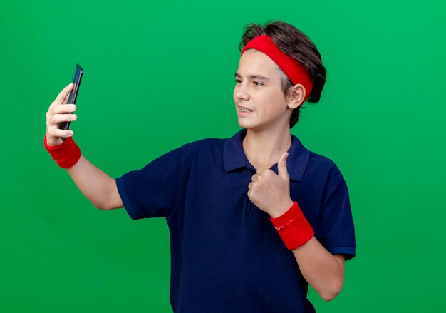 Smiling young handsome sporty boy wearing headband and wristbands with dental braces taking selfie showing thumb up isolated on green wall