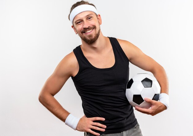 Smiling young handsome sporty man wearing headband and wristbands holding soccer ball with hand on waist isolated on white space