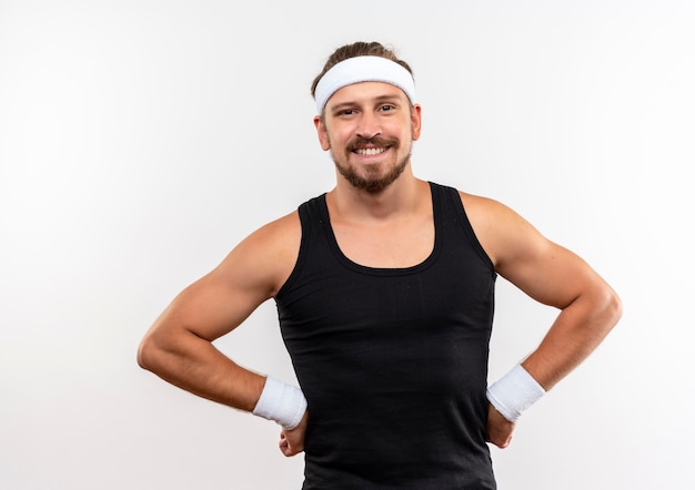 Smiling young handsome sporty man wearing headband and wristbands putting hands on waist isolated on white space