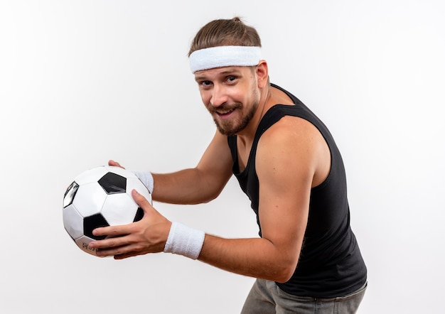 Smiling young handsome sporty man wearing headband and wristbands standing in profile view holding soccer ball isolated on white space