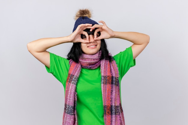 Free photo smiling young ill caucasian girl wearing winter hat and scarf  doing look gesture using hands as binoculars isolated on white wall with copy space