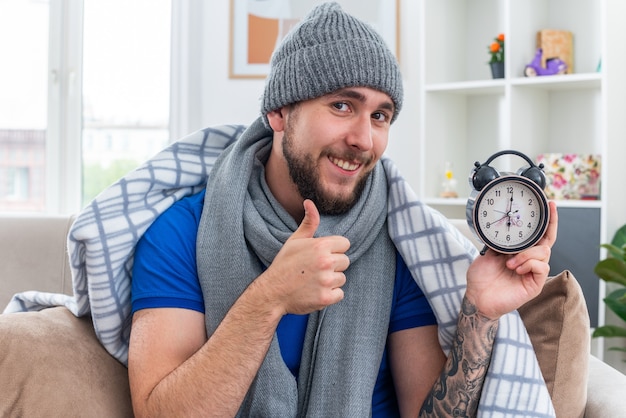 Free photo smiling young ill man wearing scarf and winter hat sitting on sofa in living room wrapped in blanket holding alarm clock  showing thumb up