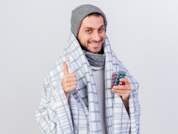 Smiling young ill man wearing winter hat and scarf wrapped in plaid holding pills