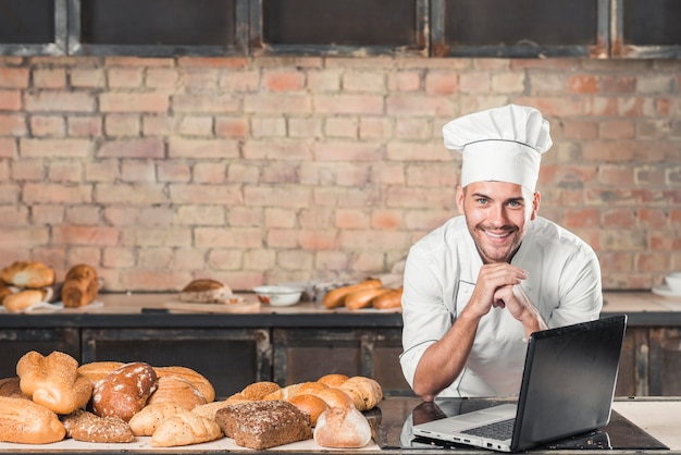 Free photo smiling young male baker leaning on table with variety of baked bread and laptop