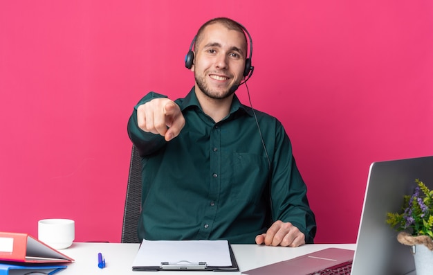 Smiling young male call center operator wearing headset sitting at desk with office tools showing you gesture 