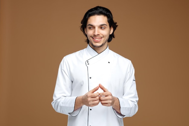 smiling young male chef wearing uniform keeping hands together looking at side isolated on brown background