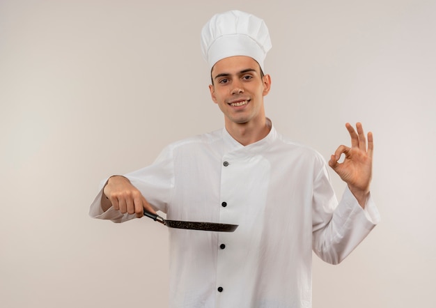 Smiling young male cook wearing chef uniform holding frying pan showing okey gesture