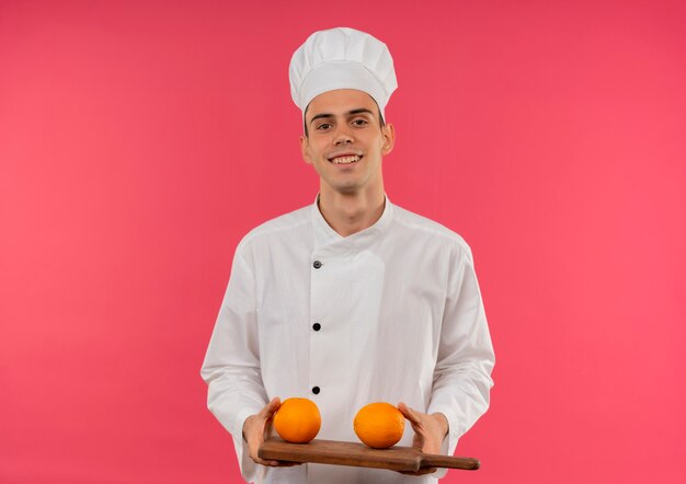 Smiling young male cook wearing chef uniform holding orange on cutting board with copy space