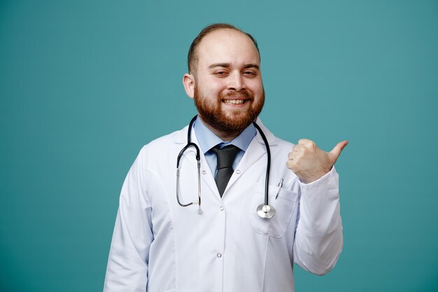 Smiling young male doctor wearing medical coat and stethoscope around his neck looking at camera pointing to side isolated on blue background