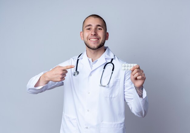 Smiling young male doctor wearing medical robe and stethoscope around his neck holding and pointing at pack of medical tablets isolated on white wall