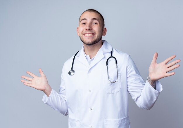 Smiling young male doctor wearing medical robe and stethoscope around his neck showing empty hands isolated on white wall