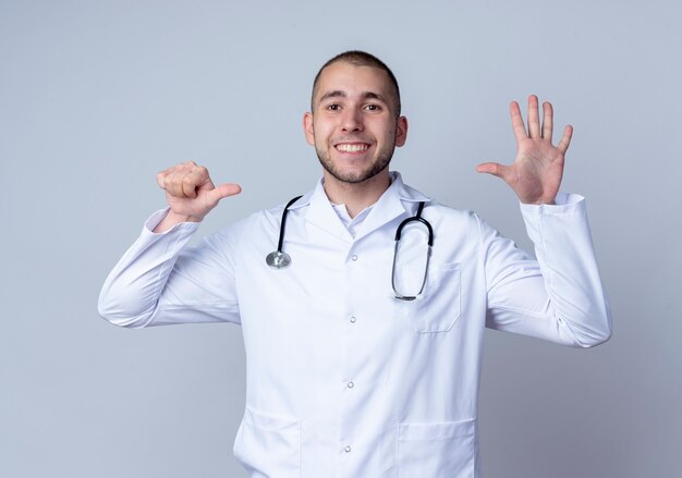 Smiling young male doctor wearing medical robe and stethoscope around his neck showing six with hands isolated on white wall