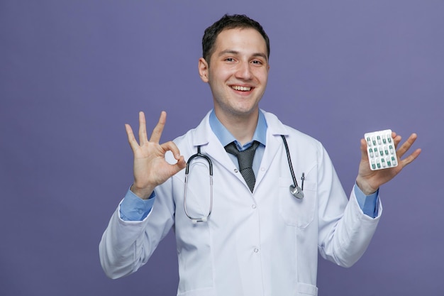 Smiling young male doctor wearing medical robe and stethoscope around neck looking at camera showing pack of capsules and ok sign isolated on purple background