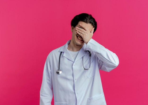 Smiling young male doctor wearing medical robe and stethoscope covering eyes with hand isolated on pink wall with copy space