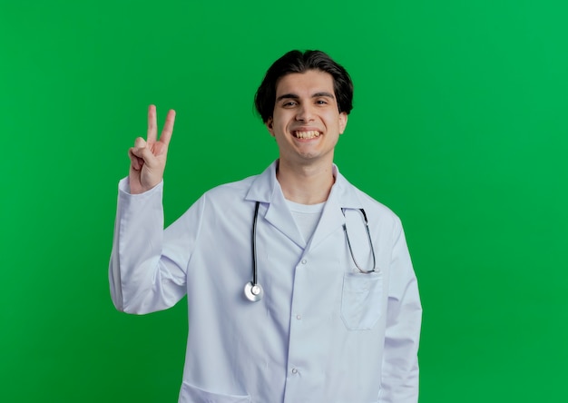 Smiling young male doctor wearing medical robe and stethoscope doing peace sign  isolated on green wall with copy space