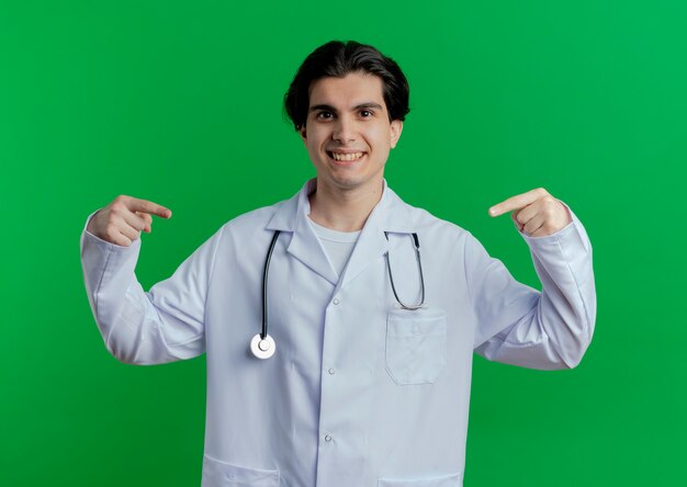 Smiling young male doctor wearing medical robe and stethoscope pointing at something in front of him isolated on green wall