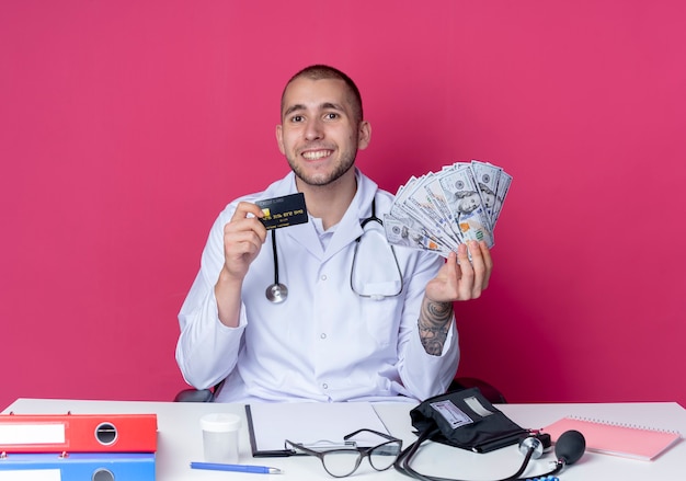 Free Photo smiling young male doctor wearing medical robe and stethoscope sitting at desk with work tools holding money and credit card isolated on pink wall