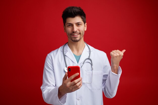 Smiling young male doctor wearing medical uniform and stethoscope around his neck holding mobile phone looking at camera pointing to side isolated on red background