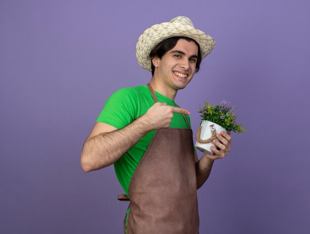 Smiling young male gardener in uniform wearing gardening hat holding and points at flower in flowerpot