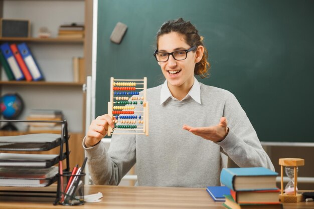 smiling young male teacher wearing glasses sitting at desk holding and points at abacus with school tools on in classroom