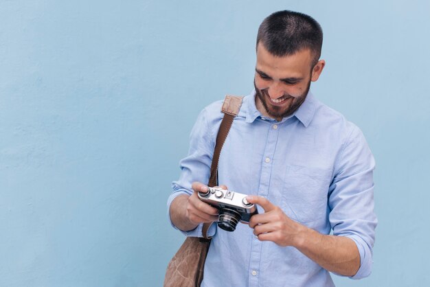 Smiling young man carrying backpack and looking at camera standing near blue wall