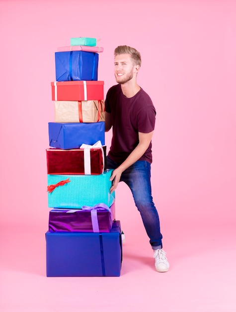 Smiling young man carrying stack of colorful presents against pink background