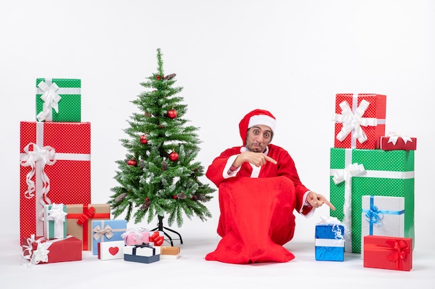 Smiling young man dressed as Santa claus with gifts and decorated Christmas tree sitting on the ground pointing down on white background