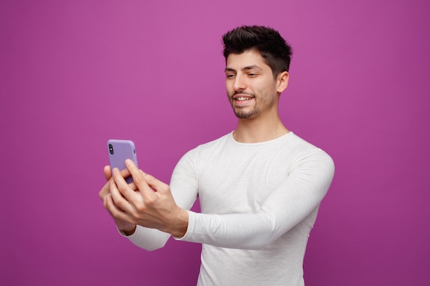 Smiling young man holding mobile phone taking selfie isolated on purple background