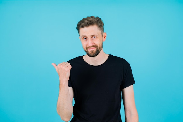 Smiling young man is pointing left with thumb on blue background