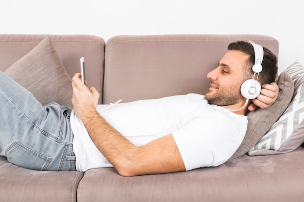 Free photo smiling young man lying on sofa listening music on headphone through mobile phone