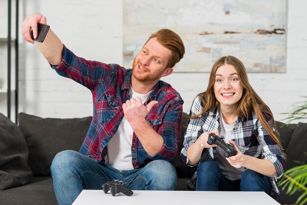 Smiling young man showing thumb up sign while taking selfie on cell phone at home