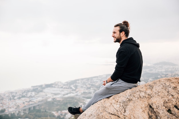 Free Photo smiling young man sitting on mountain peak looking at cityscape