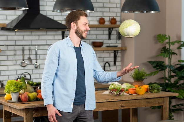 Smiling young man standing in the kitchen throwing whole cabbage in air