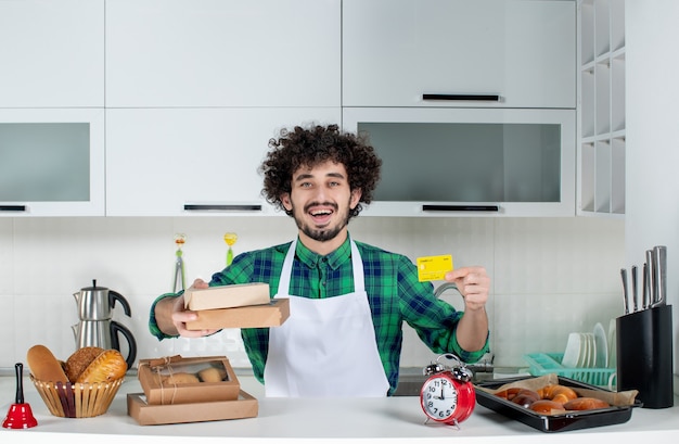 Smiling young man standing behind the table various pastries on it and holding bank card brown boxes in the white kitchen