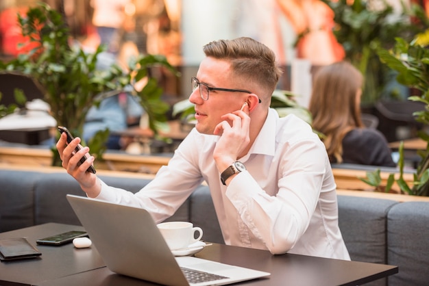 Free photo smiling young man using wireless earphone looking at mobile phone in caf�