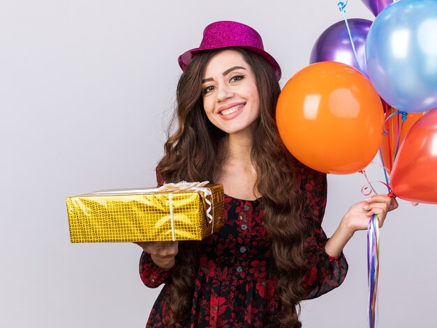 Smiling young party girl wearing party hat holding balloons and gift package looking at camera isolated on white wall